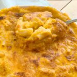 A close-up of a golden-brown baked dish, possibly Butternut Squash Macaroni and Cheese, with a spoon lifting a portion to reveal a soft, creamy interior with visible chunks, set on a light-colored surface.