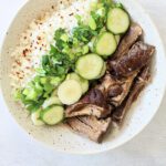 A bowl of Slow Cooker Banh Mi Bowls featuring shredded beef, sliced cucumber, chopped green onions, white rice topped with red pepper flakes, and neatly arranged sliced white vegetables.