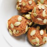A white bowl filled with round, brown pumpkin truffles topped with green pumpkin seeds, on a white background.