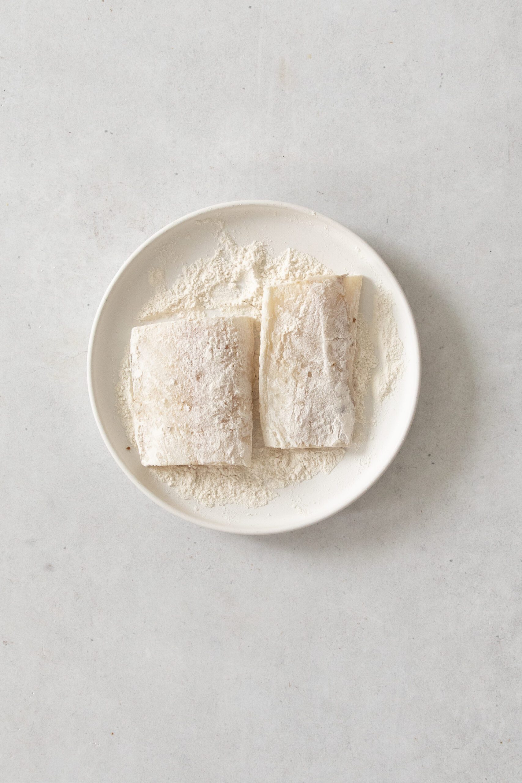 Two pieces of raw white fish coated in flour are placed on a white oval plate, with extra flour scattered around them on the plate. The background is a light gray surface.