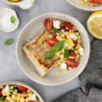 A plate with a piece of grilled fish and a side of corn salad with cherry tomatoes, feta cheese, and basil leaves, surrounded by lemon wedges and bowls of more salad on a gray surface.