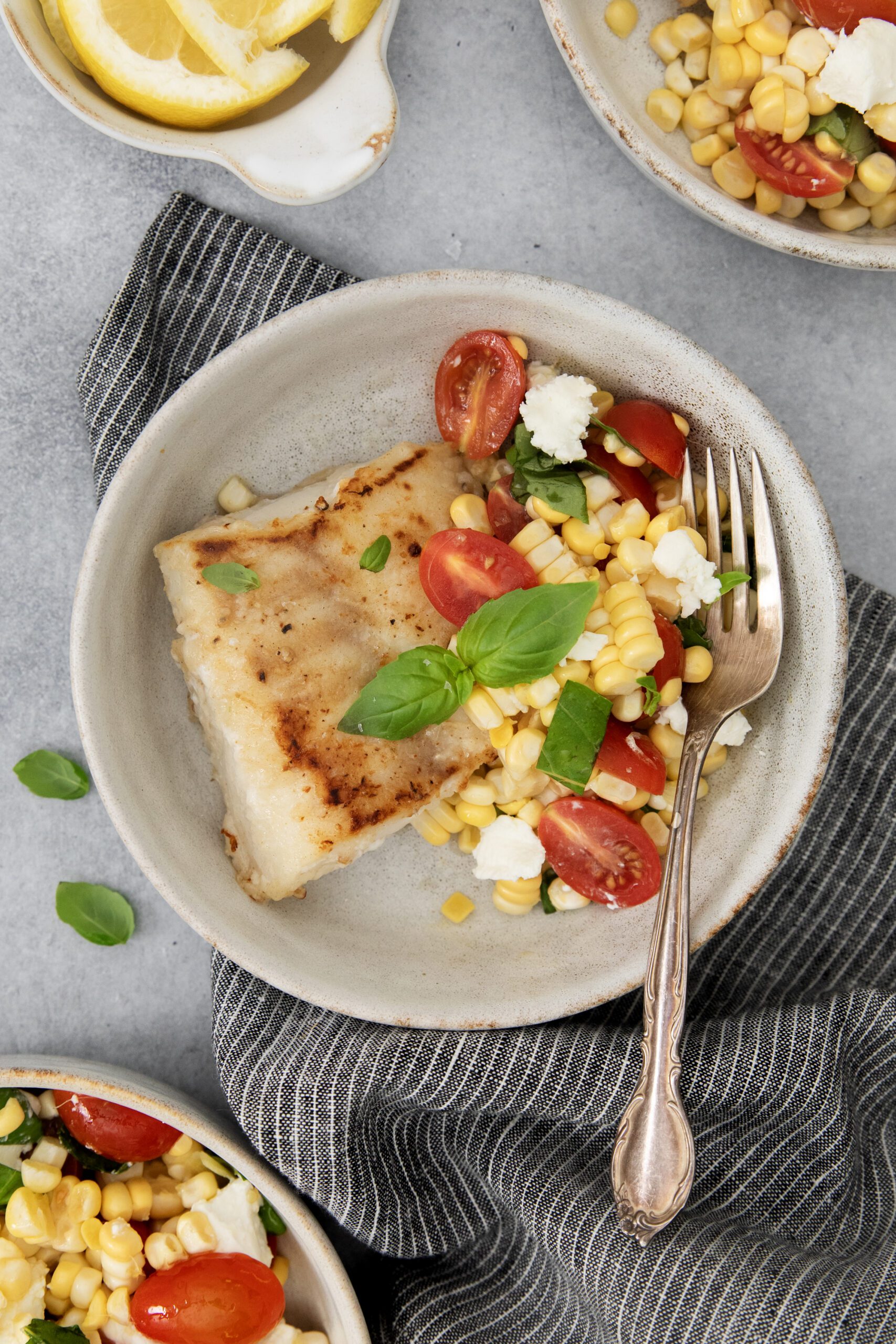 A plate with a piece of cooked fish topped with corn, cherry tomatoes, feta cheese, and fresh basil. A fork rests on the side, with lemon slices and other similar bowls nearby on a gray surface.