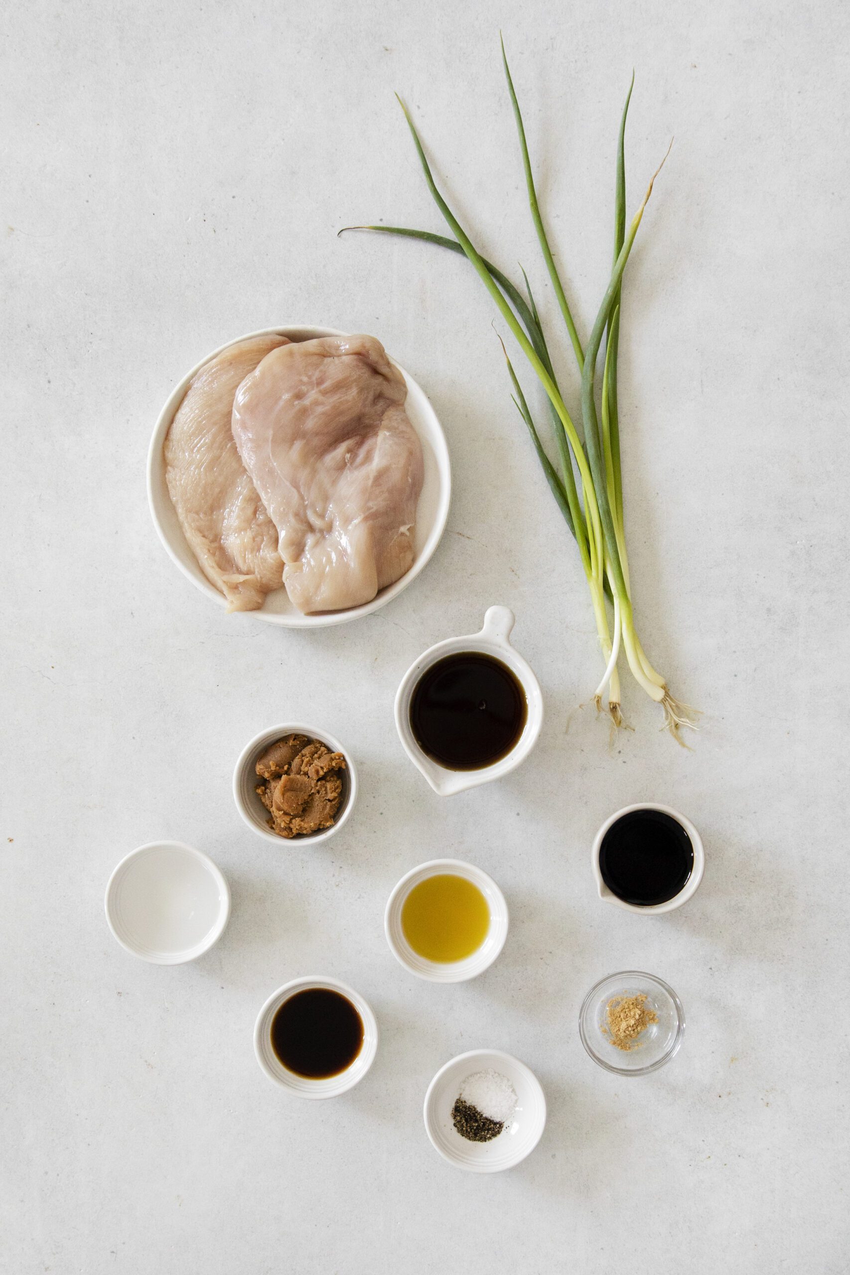 Overhead view of Miso Teriyaki Chicken ingredients: raw chicken breasts in a bowl, green onions, and small bowls with brown sugar, soy sauce, miso, oil, pepper, salt, and ginger on a light surface.