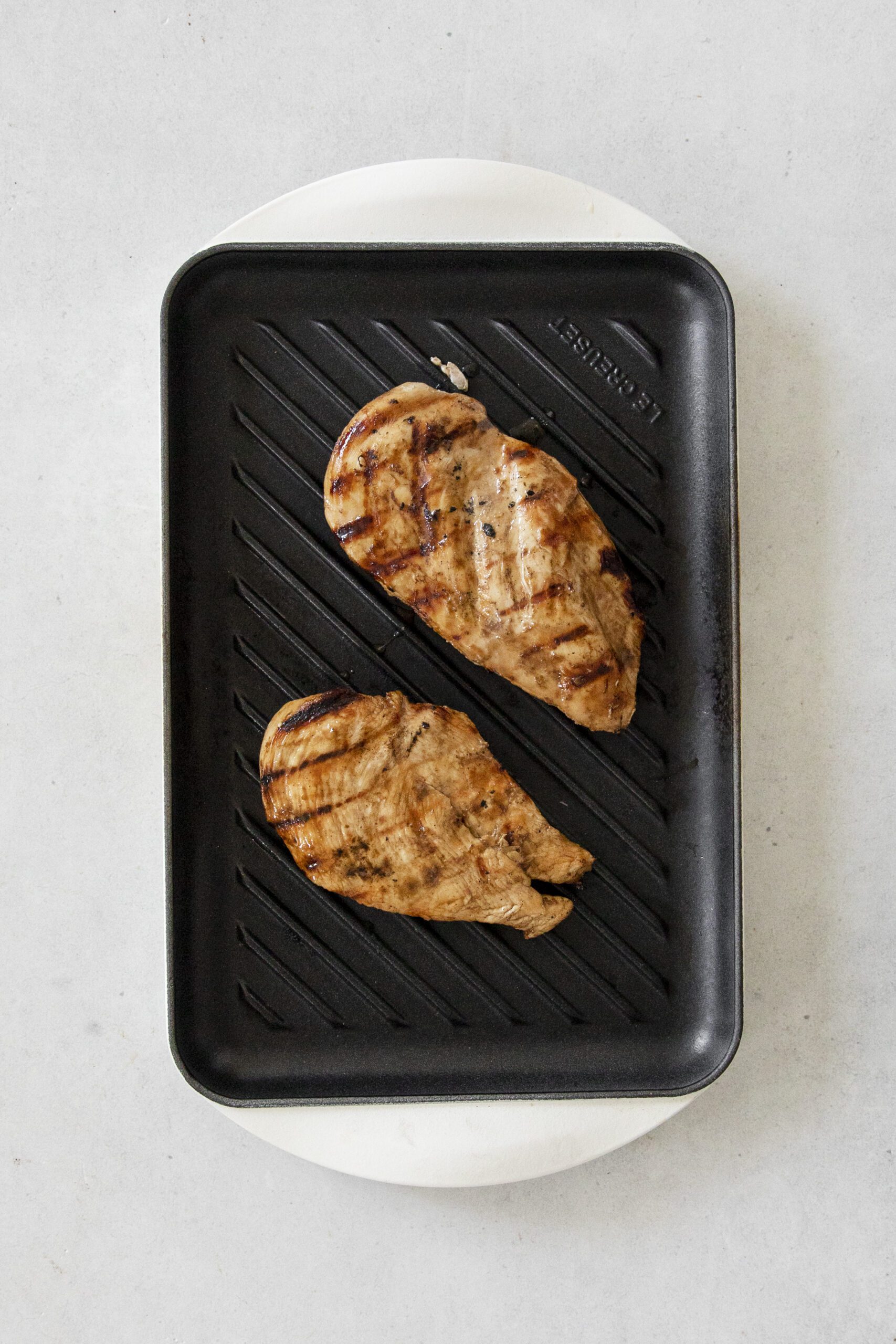 Two cooked Miso Teriyaki Chicken breasts with grill marks sit on a black grill pan atop a light-colored surface.