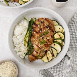 A bowl with Miso Teriyaki Chicken breast, white rice, zucchini slices, and green onions, garnished with sesame seeds. A small pitcher of sauce, a bowl of sesame seeds, and miso paste sit nearby on a checkered cloth.