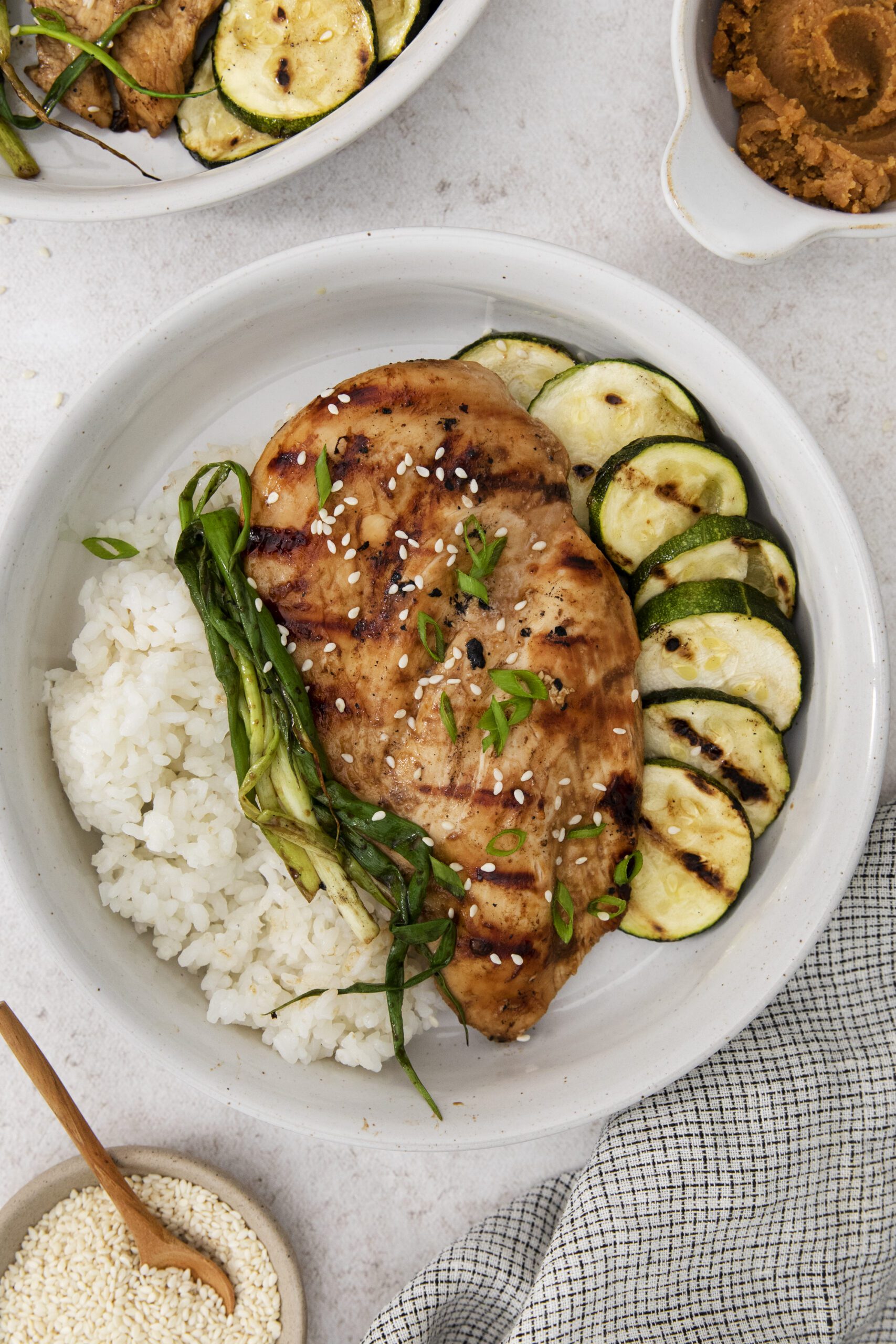 A bowl with Miso Teriyaki Chicken breast, steamed white rice, sliced grilled zucchini, and green onions, sprinkled with sesame seeds. A plate of miso paste and a bowl of sesame seeds are nearby.