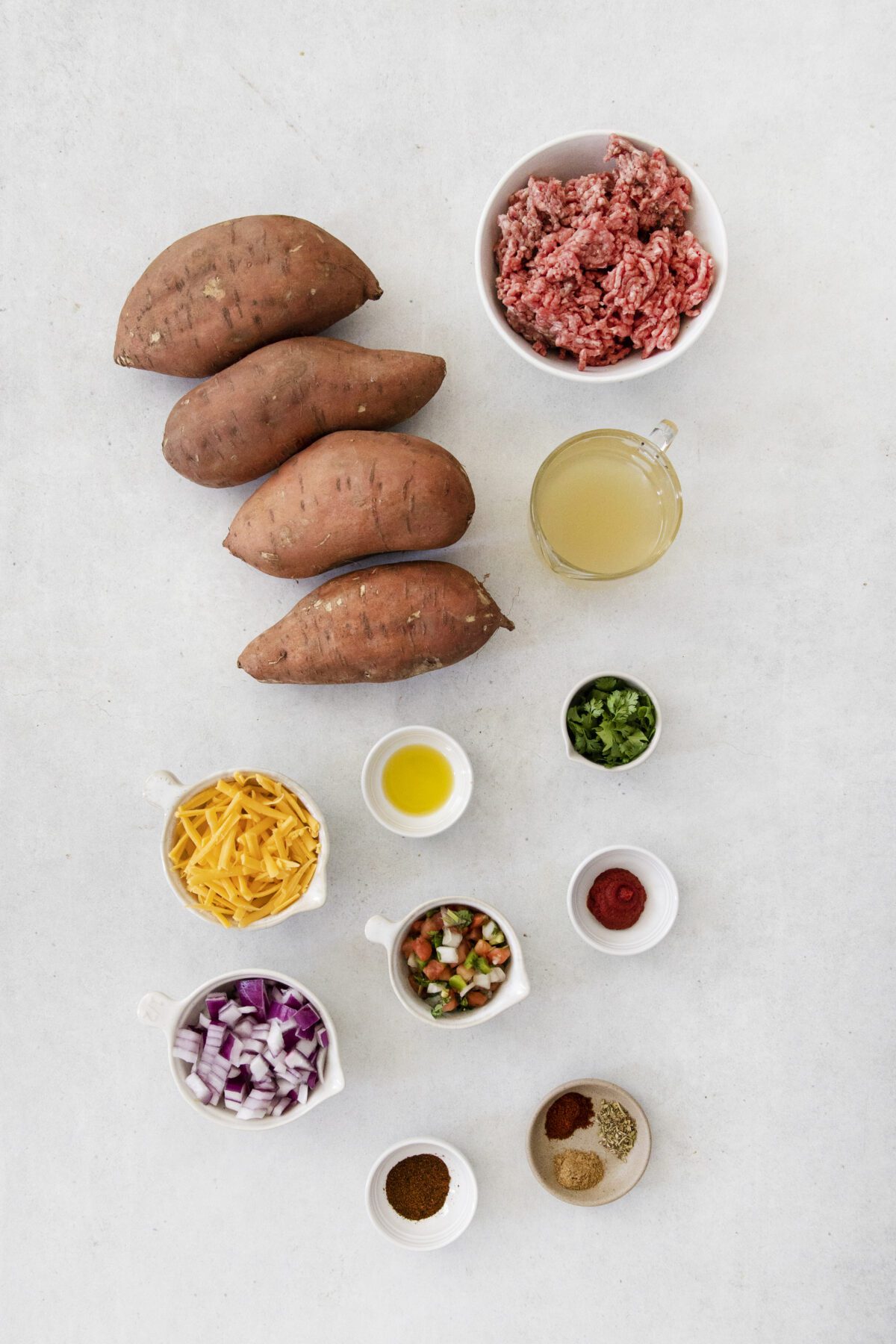 Overhead view of sweet potatoes, ground beef, broth, shredded cheese, diced red onion, pico de gallo, chopped green peppers, olive oil, ketchup, and various spices arranged neatly on a light background.