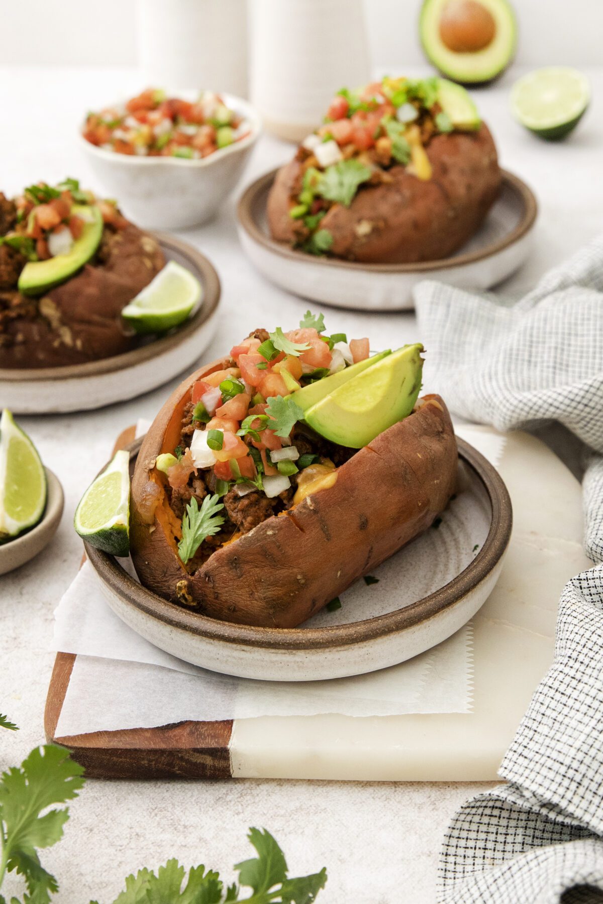 A baked sweet potato on a plate, stuffed with chili, topped with fresh salsa, avocado slices, and cilantro. Lime wedges and more stuffed sweet potatoes are in the background.