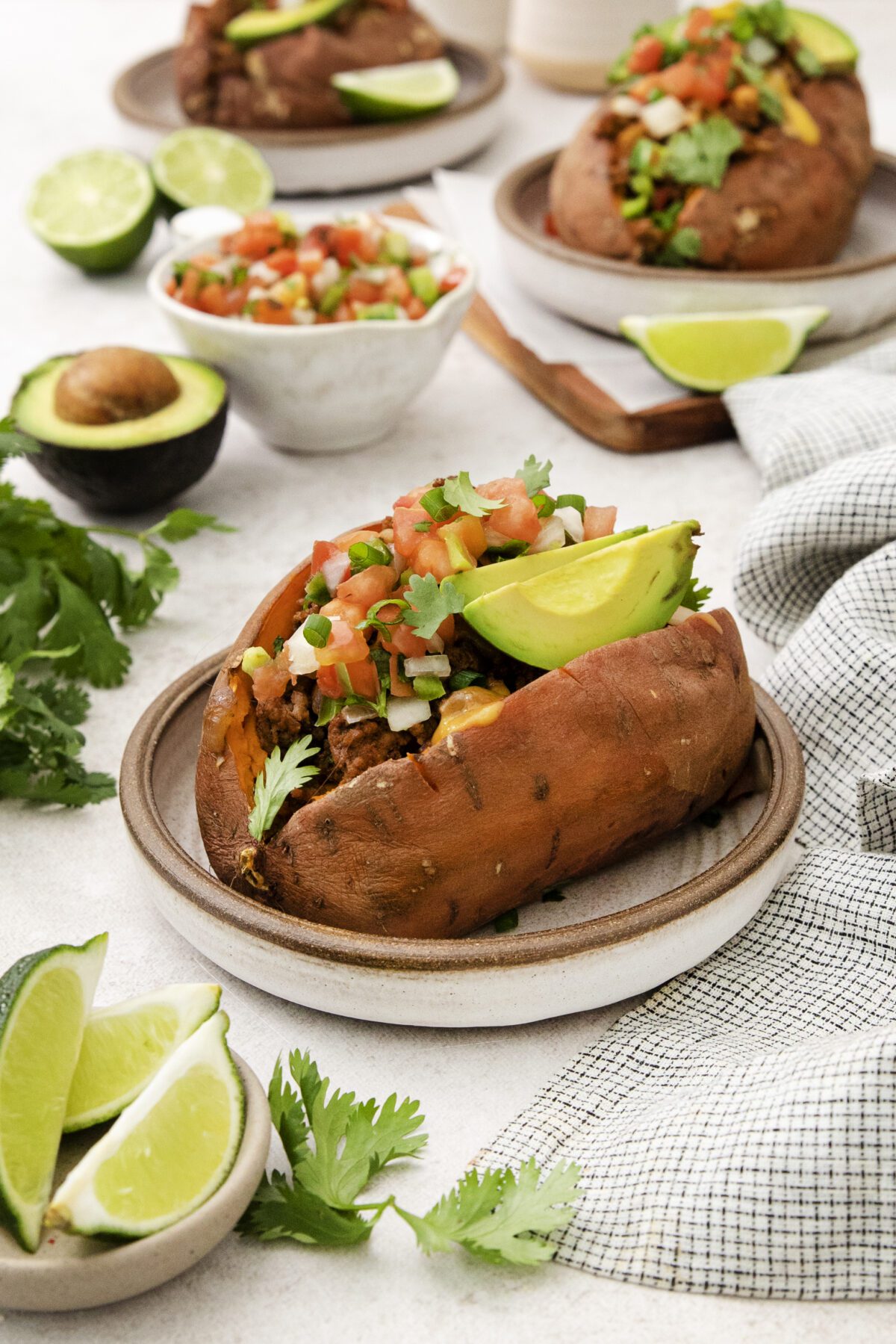 A baked sweet potato on a plate is filled with avocado slices, pico de gallo, and fresh cilantro. Surrounding it are lime wedges, an avocado, and bowls of pico de gallo, with another stuffed sweet potato in the background.