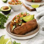 A baked sweet potato on a plate is filled with avocado slices, pico de gallo, and fresh cilantro. Surrounding it are lime wedges, an avocado, and bowls of pico de gallo, with another stuffed sweet potato in the background.