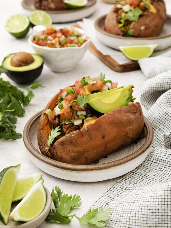 A baked sweet potato on a plate is filled with avocado slices, pico de gallo, and fresh cilantro. Surrounding it are lime wedges, an avocado, and bowls of pico de gallo, with another stuffed sweet potato in the background.