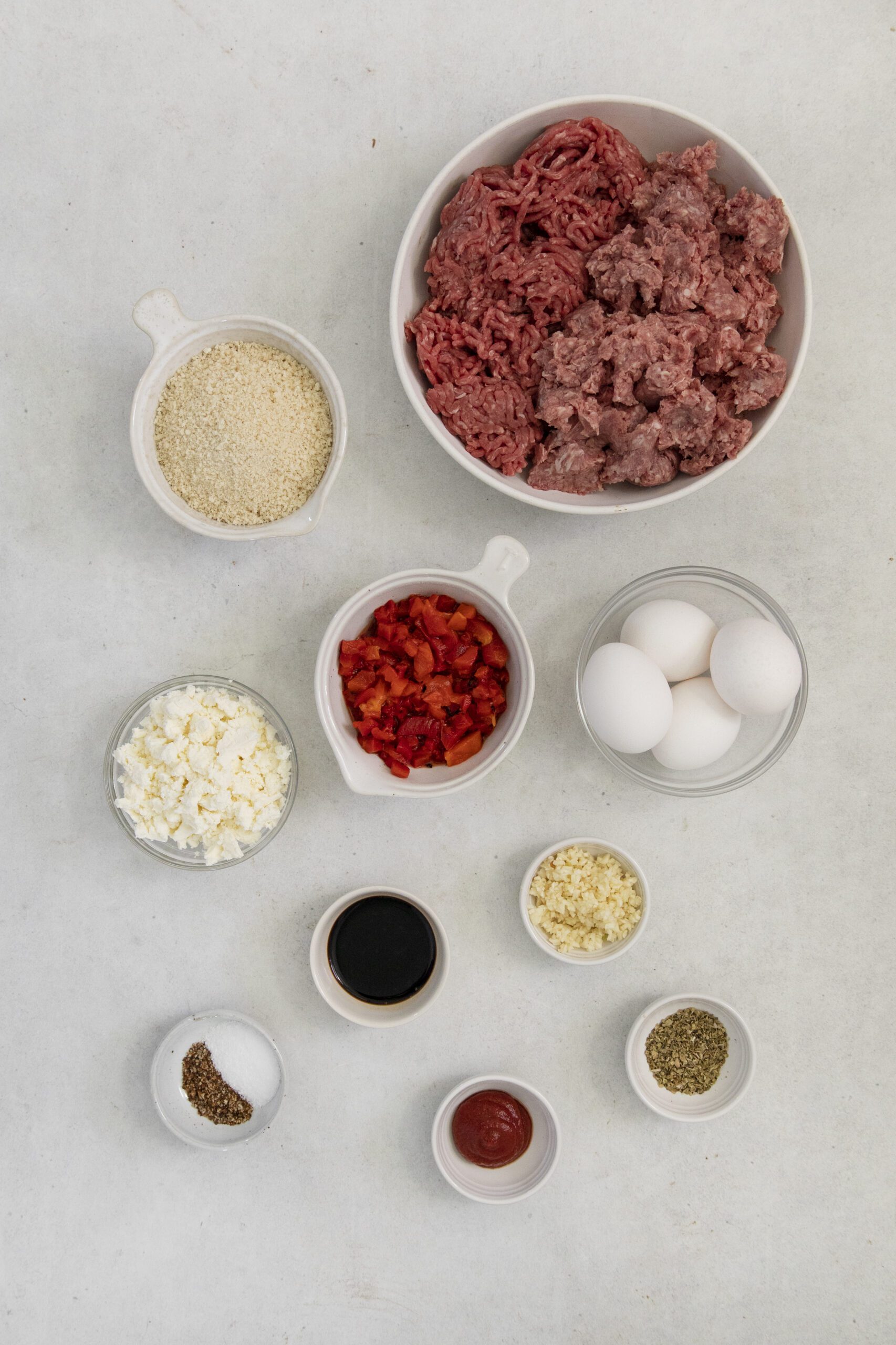 A flat lay of Greek Meatloaf ingredients in bowls on a white surface: ground meat, breadcrumbs, eggs, chopped red peppers, crumbled cheese, minced garlic, black pepper, dried herbs, sauce, and salt.