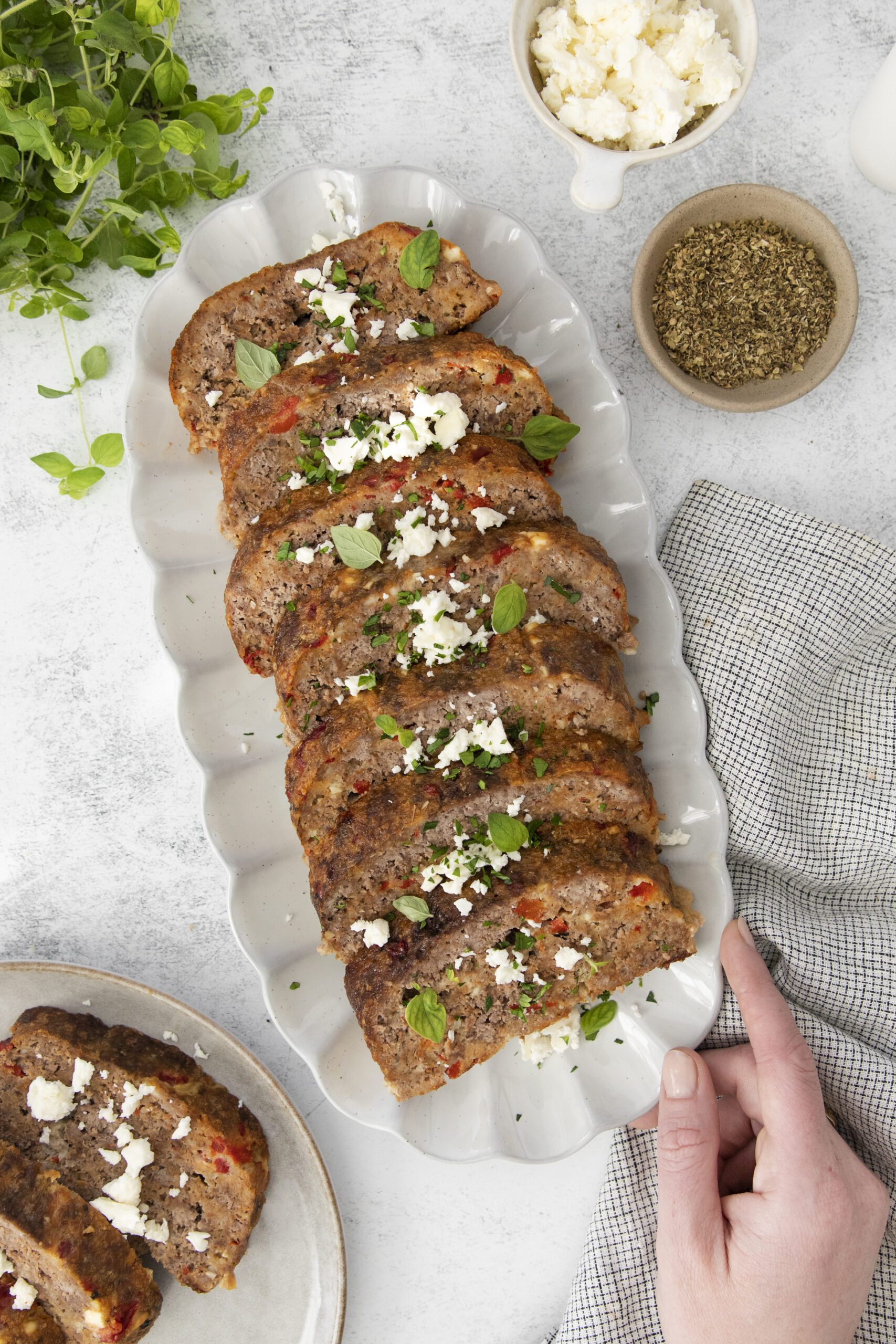 A hand reaches for a platter of Greek Meatloaf garnished with crumbled cheese and fresh herbs. A bowl of seasoning, a bowl of cheese, and a sprig of fresh herbs sit nearby on a neutral surface.