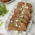 A platter of sliced meatloaf garnished with crumbled cheese and fresh herbs, surrounded by herbs, a bowl of cheese, and a checkered napkin on a light-colored surface.