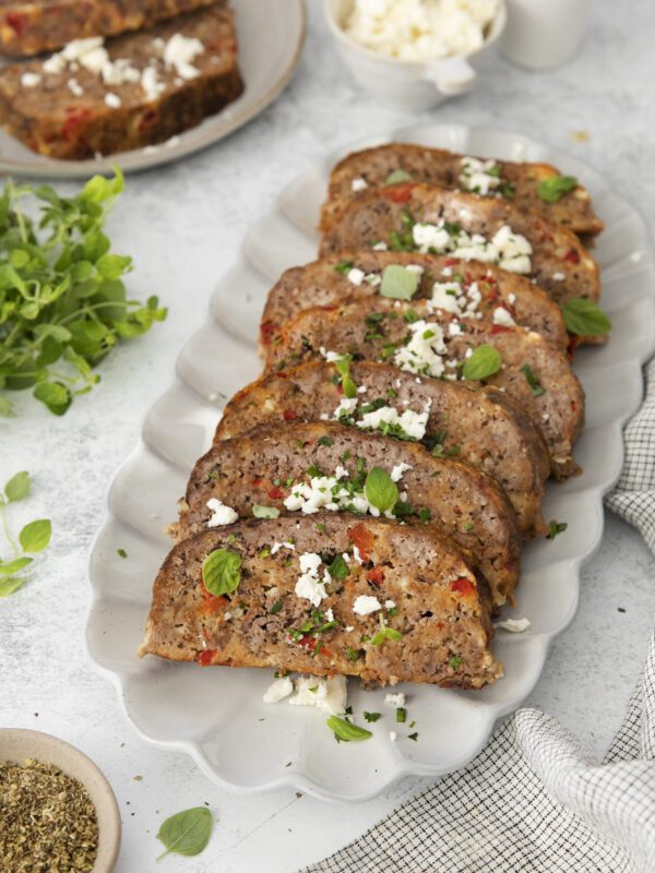 A platter of sliced meatloaf garnished with crumbled cheese and fresh herbs, surrounded by herbs, a bowl of cheese, and a checkered napkin on a light-colored surface.