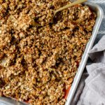 A baking tray filled with golden-brown homemade granola sits on a gray striped cloth. A gold spoon rests in the granola, ready for serving.