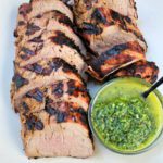 Sliced Citrus Pork tenderloin arranged on a plate beside a small bowl of green chimichurri sauce with a spoon.