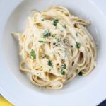 A plate of creamy Cauliflower Alfredo pasta garnished with grated cheese and fresh herbs sits on a white dish. Two lemons rest nearby on a blue textured cloth in the background.