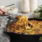 A hand lifts a cheesy serving of zucchini lasagna recipe from a skillet with a fork; melted cheese stretches from the pan, and fresh herbs garnish the top. Dishes, vases, and herbs decorate the background.