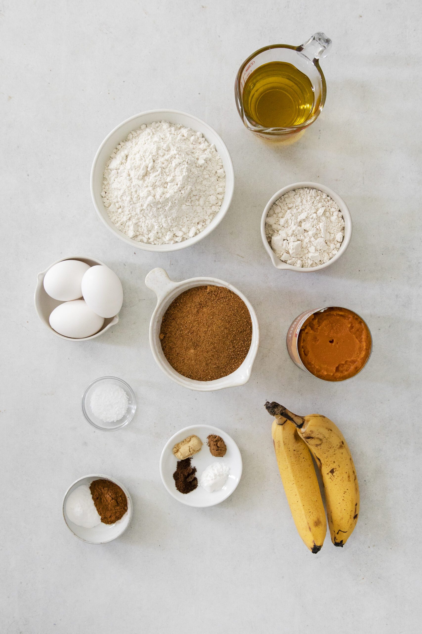 A top-down view of baking ingredients arranged on a white surface, perfect for making Pumpkin Banana bundt cake—featuring flour, eggs, sugar, oil, spices, baking powder, baking soda, salt, and two ripe bananas.