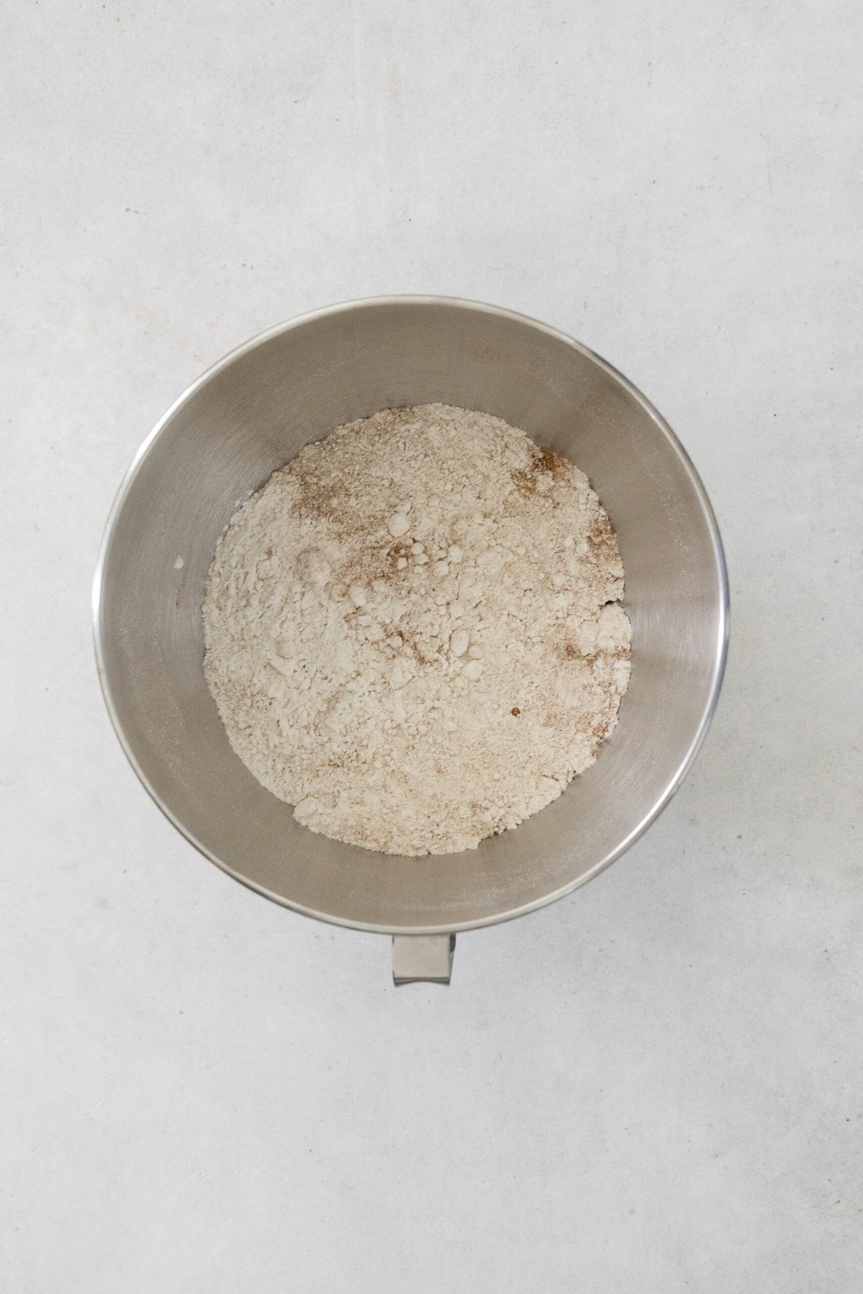 A metal mixing bowl containing a blend of dry ingredients for Pumpkin Banana bundt cake, including flour and spices, sits on a light gray surface.