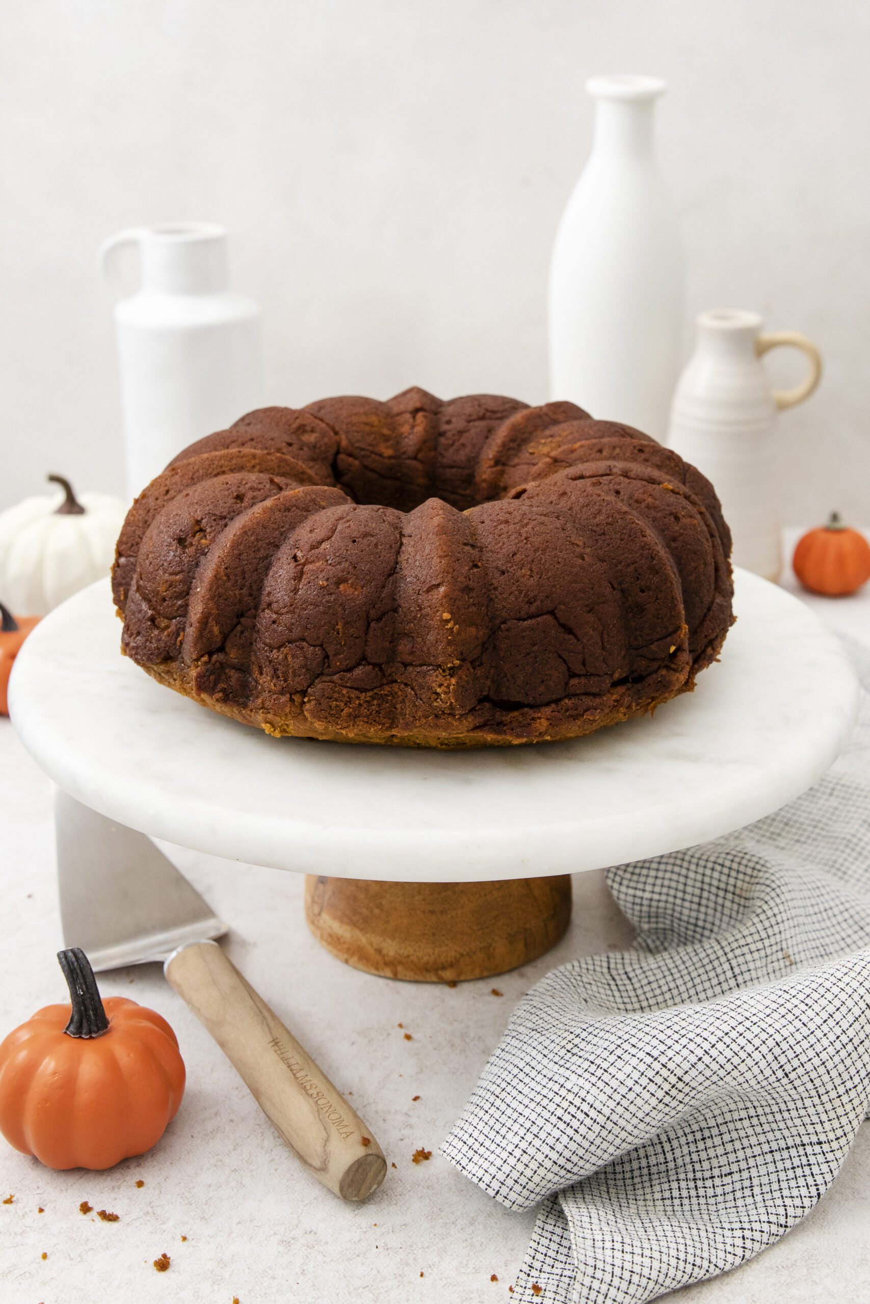 A round, golden-brown Pumpkin Banana bundt cake sits on a white marble cake stand, surrounded by small decorative pumpkins and ceramic pitchers on a light table with a checkered cloth nearby.