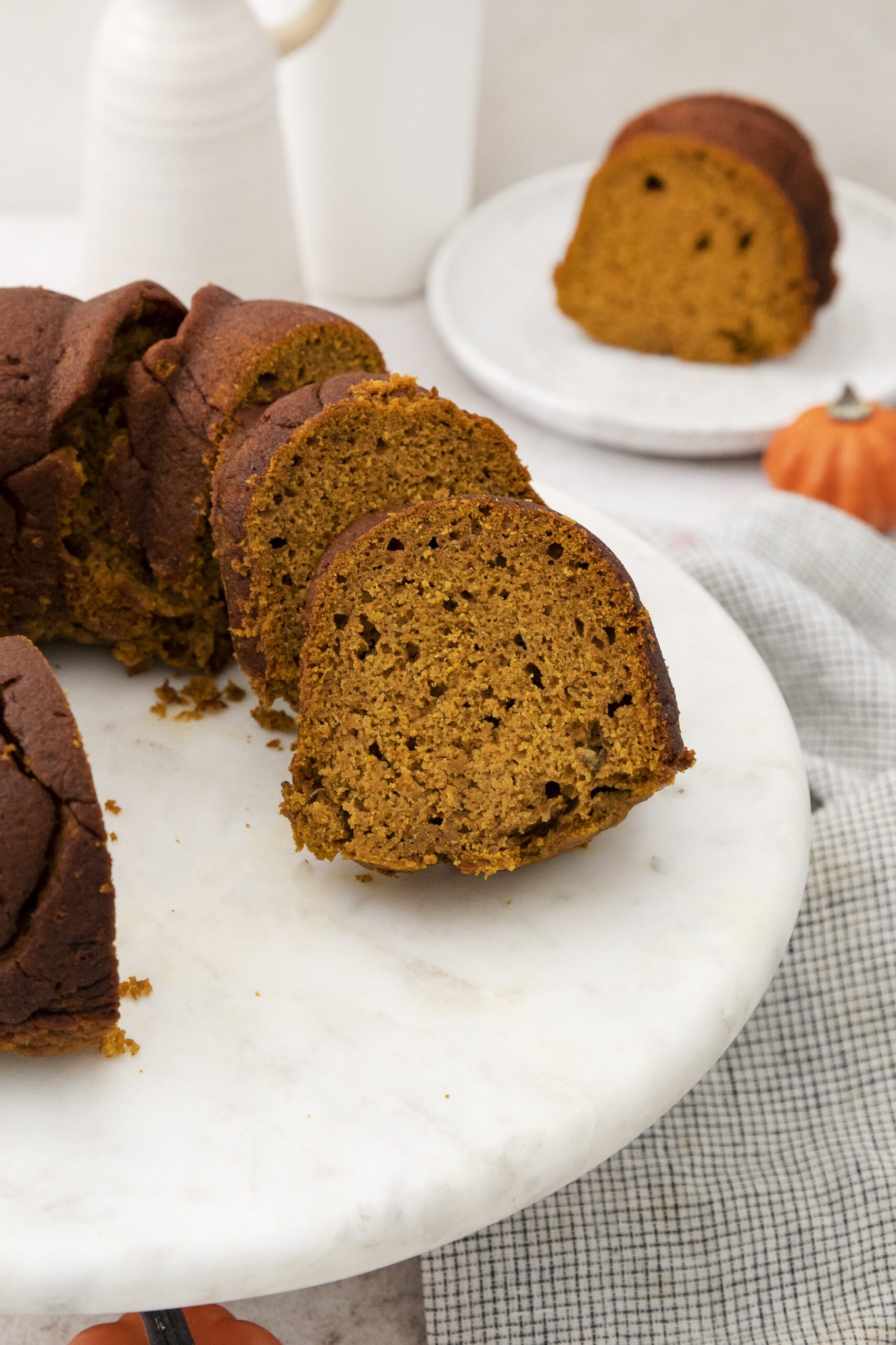 A sliced Pumpkin Banana bundt cake is displayed on a marble cake stand, with a few slices arranged in front. A small pumpkin and a plate with an additional cake slice are in the background.