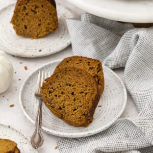 Two slices of Pumpkin Banana bundt cake sit on a speckled ceramic plate with a fork. More cake slices and a whole bundt cake are visible nearby, along with a white napkin and small decorative pumpkins.