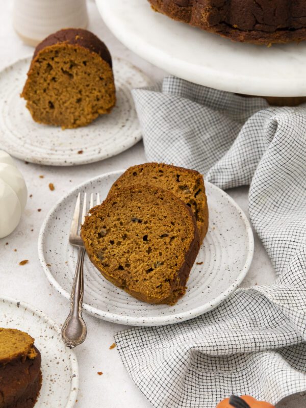 Two slices of Pumpkin Banana bundt cake sit on a speckled ceramic plate with a fork. More cake slices and a whole bundt cake are visible nearby, along with a white napkin and small decorative pumpkins.