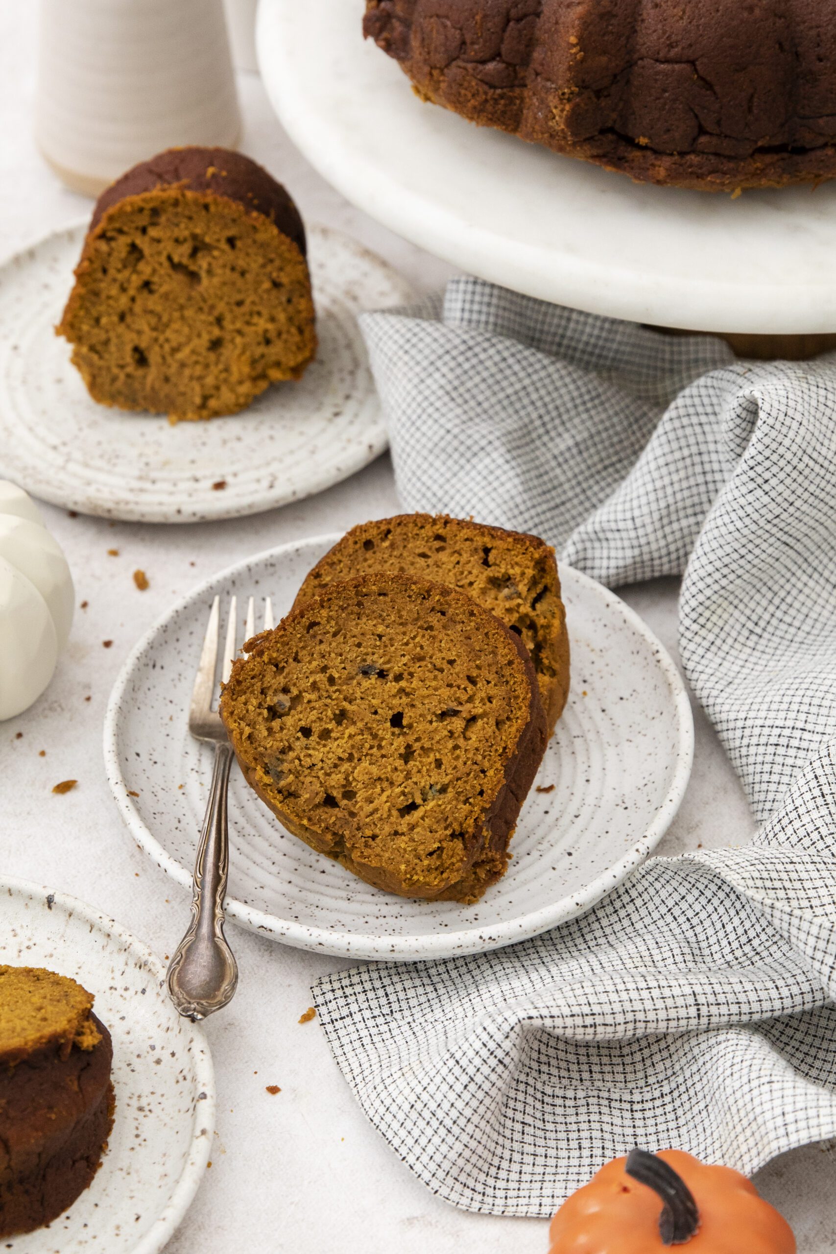 Two slices of Pumpkin Banana bundt cake sit on a speckled ceramic plate with a fork. More cake slices and a whole bundt cake are visible nearby, along with a white napkin and small decorative pumpkins.