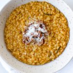 A bowl of creamy orange-colored risotto garnished with pecans and grated cheese, served in a speckled ceramic dish on a white surface.