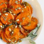 A white plate with sliced roasted sweet potatoes topped with green pumpkin seeds, drizzled sauce, and garnished with fresh sage leaves—a vibrant roasted sweet potato recipe for any occasion.