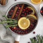A simmer pot filled with cranberries, orange slices, rosemary sprigs, and cinnamon sticks sits on a light surface. Extra ingredients and a striped cloth are arranged nearby, ready to create a cozy, inviting aroma.