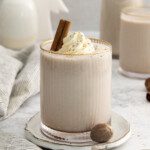 A glass of creamy vegan eggnog topped with whipped cream and a cinnamon stick sits on a plate, with whole nutmeg nearby. A second glass and decorative objects are blurred in the background.