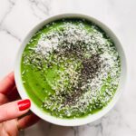 A hand with red nail polish holds a vibrant smoothie bowl filled with green smoothie, topped with shredded coconut, chia seeds, and hemp seeds on a white marble surface.