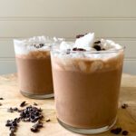 Two glasses of creamy chocolate smoothie topped with whipped cream and cacao nibs sit on a wooden surface, with extra cacao nibs scattered around. The delicious smoothie is set against a light-colored, paneled wall in the background.