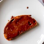 A slice of bread with Nutella chocolate spread on top sits on a white plate, with a small smear and knife visible at the edge of the image.