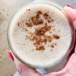 A hand with red-painted nails holds a glass of frothy smoothie topped with sprinkled cinnamon, viewed from above against a light wood floor background.