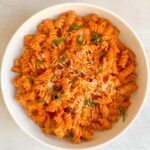 A white bowl filled with Spicy Tomato Pasta—rotini in a creamy, zesty tomato sauce, topped with grated cheese and fresh basil—placed on a light-colored surface.