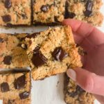A hand holds a square bar with a golden, crumbly texture and large chocolate chunks—one of 5 Healthier Dessert Recipes; other similar bars rest on a white surface below.