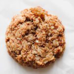 A close-up of a single oatmeal raisin breakfast cookie with visible oats and a rough texture, placed on a white surface.