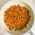 A white frying pan filled with spiced chickpeas coated in reddish-brown spices, placed on a light-colored surface.