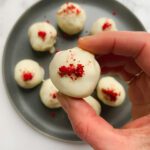 A hand holds a strawberry shortcake truffle topped with red crumbs above a dark plate filled with more strawberry shortcake truffles, all garnished with red sprinkles, on a white surface.