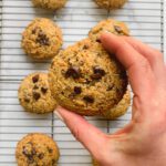 A hand holds a grain free chocolate chip cookie above a cooling rack with several more cookies spread out in the background.