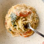 A fork twirls creamy one-pan spaghetti with spinach, tomatoes, and grated cheese in a speckled beige bowl.
