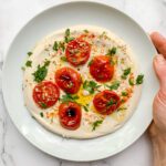 A white plate with creamy hummus topped with roasted cherry tomatoes, chopped parsley, sesame seeds, olive oil, and spices, held by a hand over a white marble surface.