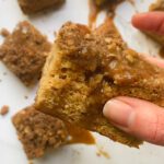 A close-up of a hand holding a square piece of crumb-topped salted caramel coffee cake, with more pieces and drizzled caramel visible on a white surface in the background.