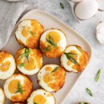 A platter of Bacon and Egg Cups topped with melted cheese and sliced green onions, surrounded by a carton of eggs, a bowl of salt, a striped napkin, and a small dish of extra green onions.