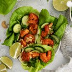 Two spicy shrimp lettuce cups filled with cucumber slices, chopped cashews, and fresh herbs are served with lime wedges on a white plate. Cashews, lime, and greens are artfully arranged around the plate.