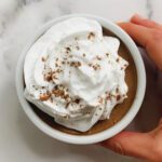 A hand holds a small white bowl filled with vegan chocolate mousse, topped with swirls of whipped cream and sprinkled with cocoa powder, on a white marble surface.